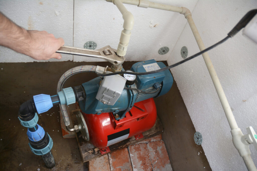 Plumber using a wrench on a water well jet pump connected to a pressure tank and water pipes.